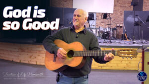 Piet Uys joyfully playing an acoustic guitar on the Fountain of Life church stage, with the bold text "God is so Good" displayed on the left side of the screen.