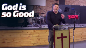 JohnDaniel speaking from behind a podium featuring a red cross. The text "God is so Good" is displayed prominently next to him against the backdrop of a church stage.
