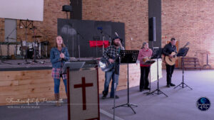 A worship team leading an unplugged, acoustic worship session on a dimly lit church stage during a power outage, featuring a vocalist, two acoustic guitarists, and a percussionist playing hand drums.