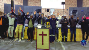 A large group of men from the HDT Academy joyfully singing, smiling, and worshipping together on the church stage, with several men raising their hands in praise.