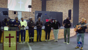 A group of men from the HDT Academy standing together on the church stage, singing in worship while Piet plays an acoustic guitar alongside them.
